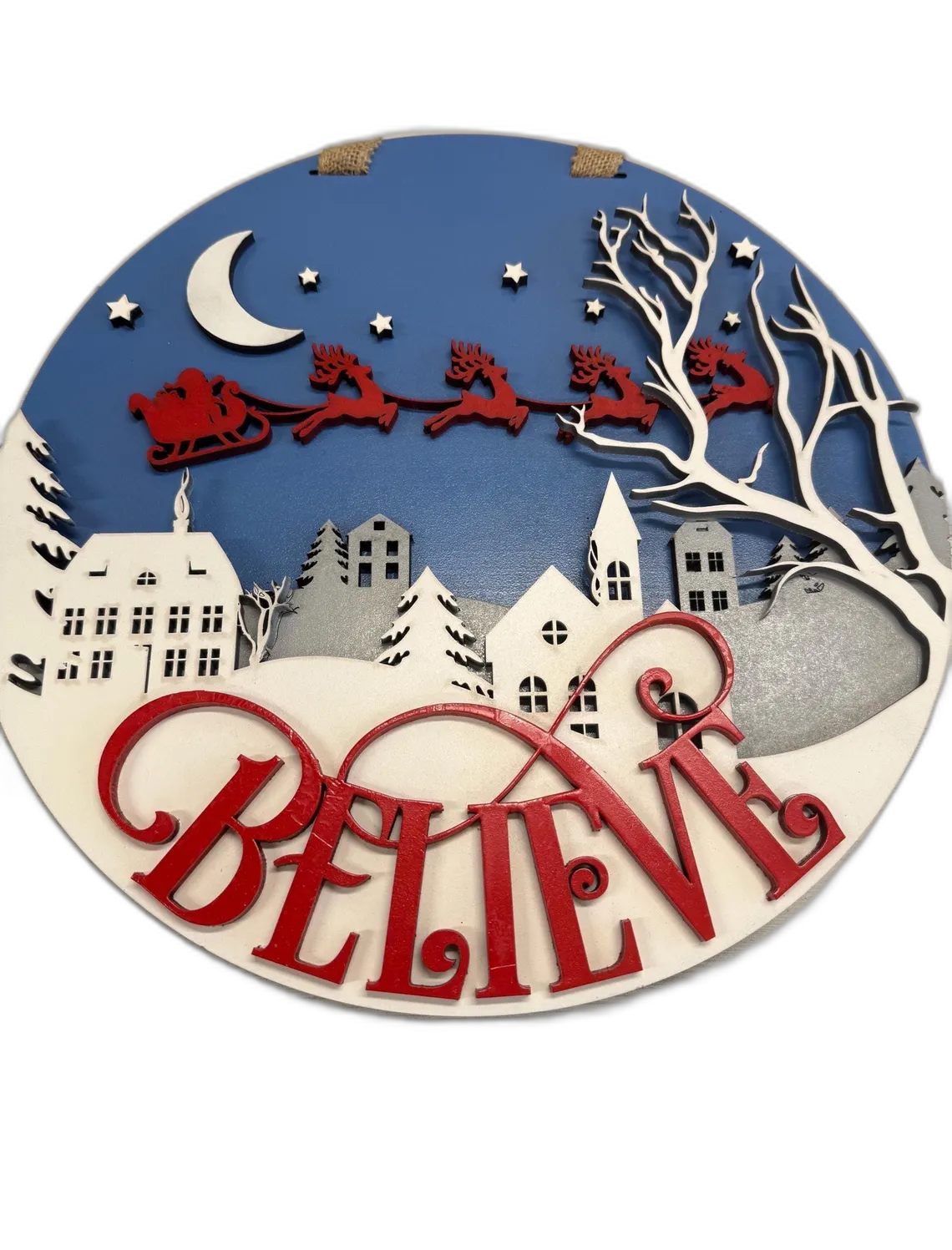 Round wood sign with Santa's sleigh flying over a snowy village and 'Believe' lettering